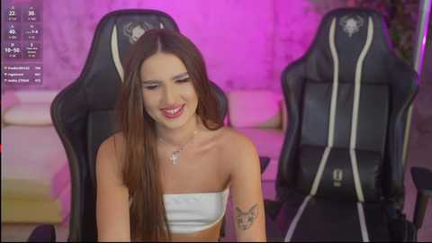 Video of a smiling, light-skinned woman with long brown hair in a white strapless top, sitting in a black gaming chair. Background features a purple-lit room with a white couch.