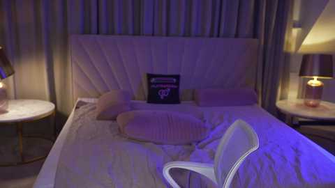 Video of a dimly lit, modern hotel room with a plush white headboard, beige sheets, and a white chair. Nightstands with lamps flank the bed, and a framed picture hangs above it.