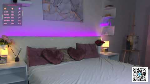 A video of a modern bedroom with a white bed adorned with purple pillows and a heart-shaped pillow. The room has a minimalist aesthetic, featuring a purple LED strip light and contemporary decor.