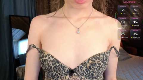 A video of a fair-skinned woman with a slender physique, wearing a leopard-print off-the-shoulder dress, standing in a bedroom with a bed and curtains visible in the background.