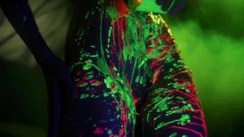A close-up video of a person's thighs covered in vibrant, neon green and red paint, creating a dynamic, abstract pattern against a dark, blurred background.