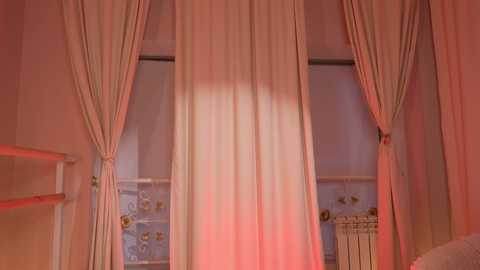 Video of a window with soft, flowing white curtains and subtle red lighting, revealing a glimpse of a decorative radiator and floral wallpaper in the background.