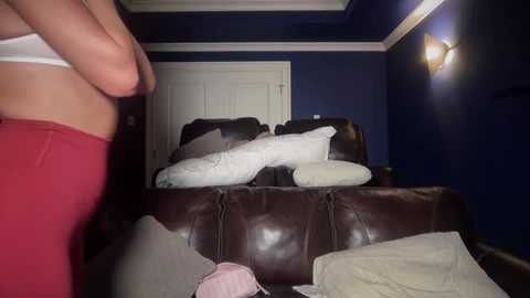 Video of a messy living room with a woman in a red top and pink bra, surrounded by a brown leather couch with scattered white pillows and blankets, against dark blue walls.