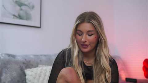 Video of a smiling, light-skinned blonde woman with long hair, wearing black lingerie, sitting on a gray tufted sofa, with a framed floral painting and a red lamp in the background.