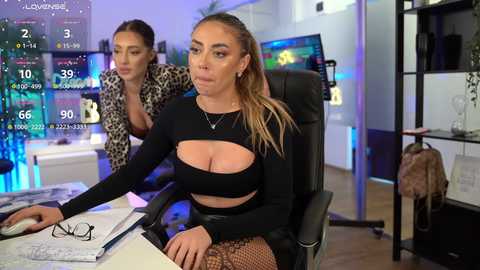 Video of two women in a modern office: one blonde, large-breasted, wearing a black crop top and fishnet tights, sits at a desk; another brunette in a leopard print jacket stands behind her.