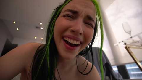 Video of a young woman with long, bright green hair, wearing a black tank top, laughing joyfully, with a nose ring, in a modern, dimly-lit room with recessed lights and a white ceiling fan.