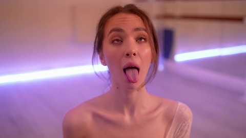 A video of a young woman with fair skin, brown hair, and a light pink top, sticking out her tongue playfully. She is in a brightly lit, modern room with purple and white lighting.