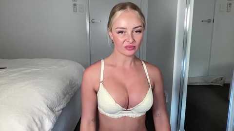 Media: Video of a blonde woman with fair skin, medium build, wearing a pale yellow lace bra, sitting in a minimalist bedroom with white walls and a bed.
