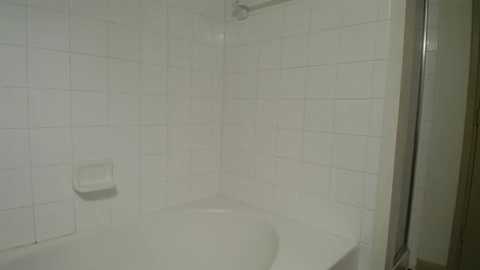 Video of a minimalist, white-tiled bathroom with a small, rectangular bathtub in the center. The walls are covered in square, white tiles, and a metal shower rod is visible in the upper right corner. The scene is clean and utilitarian.