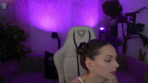 A video of a young woman with light skin and brown hair styled in a bun, wearing a white top, seated on a gaming chair. The room is dimly lit with purple lighting, featuring a plant and a computer monitor in the background.