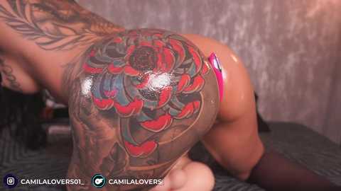 Video of a tattooed, nude woman on all fours, showcasing intricate floral and red heart designs on her back and buttocks. The background is a textured, muted grey wall.