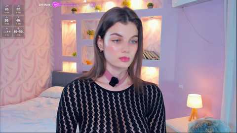 Media: A video of a young woman with fair skin, brown hair, and pink lipstick, wearing a black sheer top, standing in a pastel-colored bedroom with a bed, wall shelves, and a lamp.