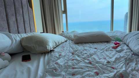 Video of a modern, minimalist bedroom with a large window showing a serene blue ocean view. The room features two twin beds with white sheets and floral pillows, a smartphone on the bedside table, and a pink bra on the floor.