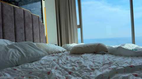 Video of a modern, well-lit bedroom with a large window showcasing a clear blue sky and distant ocean view. The bed features white sheets with a subtle floral pattern, surrounded by white pillows. The headboard is upholstered in a textured, mauve fabric.