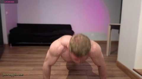 Video of a muscular, shirtless man with short blonde hair performing a push-up on a hardwood floor, in a minimalist room with a black couch and wooden table.