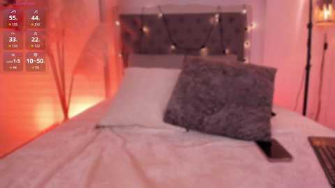 Video of a cozy bedroom with a gray tufted headboard, white bed linens, and a plush gray pillow. Warm orange lighting creates a soft ambiance. Smartphone and remote control on the bed.