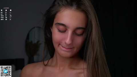 Video of a young woman with long, straight brown hair and fair skin, smiling softly, topless, in a dimly lit room with a fan and a blurred background.
