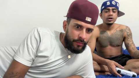 Video of two men, one in a white shirt and maroon cap, the other shirtless with tattoos, both looking at a video game controller.