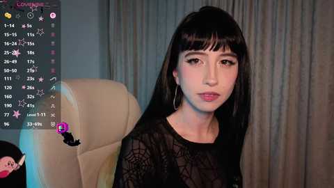 Media: Video of a pale-skinned woman with long black hair, wearing a sheer black lace top, sitting on a cream leather chair. The background features a gray curtain.