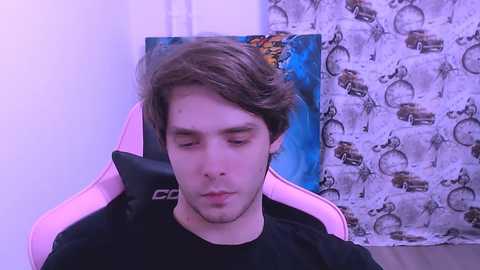 A video of a young man with light skin and brown hair sitting in a pink gaming chair, wearing a black shirt. Background includes a blue and white patterned wall with cars and a light-colored curtain.