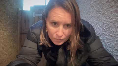 Video of a woman with shoulder-length blonde hair in a black puffer jacket, leaning forward, looking serious. Background shows a rough wooden wall and gravel ground.