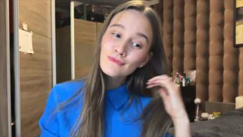 Video of a young woman with long, straight blonde hair, wearing a blue shirt, smiling, touching her hair, in a cozy, dimly-lit bedroom with a tufted headboard and cluttered nightstand.