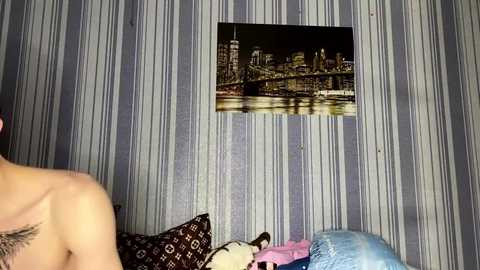 Video of a man with a tattooed shoulder, sitting on a bed with striped wallpaper and a framed New York City skyline photo.