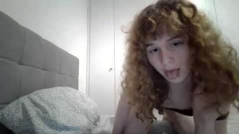Video of a young woman with curly red hair, pale skin, and a playful expression, leaning on a bed with a grey headboard and white walls.