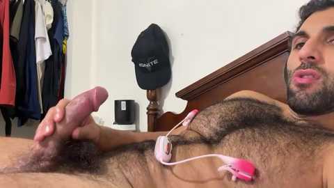 Video of a muscular, hairy, bearded man with a circumcised penis, lying naked on a bed, holding his erect penis. Background features a closet with clothes, a black hat, and a white mug.