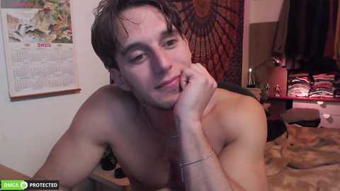 Video of a shirtless, fair-skinned man with wet hair, leaning on his elbow, smiling, in a cozy bedroom with a calendar and a colorful tapestry in the background.