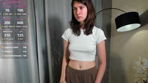 Video of a young Caucasian woman with shoulder-length brown hair, wearing a white crop top and brown pants, standing in a modern room with a digital clock, lamp, and floral decor.