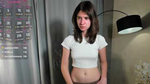 Video of a young woman with fair skin, straight dark brown hair, and medium build, wearing a tight white crop top. Background features a wall calendar, lamp, and beige curtains.