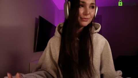 Video of a smiling Asian woman with long black hair, wearing beige hoodie, large headphones, dimly lit room with purple lighting, flat-screen TV in background.