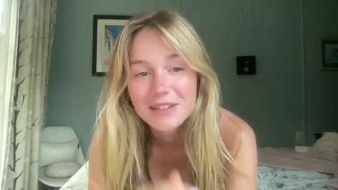 A video of a blonde, fair-skinned woman, topless, smiling, in a dimly lit, teal-painted bedroom with white furniture and framed artwork on the wall.