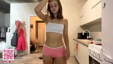 Video of a young Caucasian woman with shoulder-length brown hair, wearing a white bandeau top and pink shorts, standing in a messy kitchen with a stove, cabinets, and hanging coats.