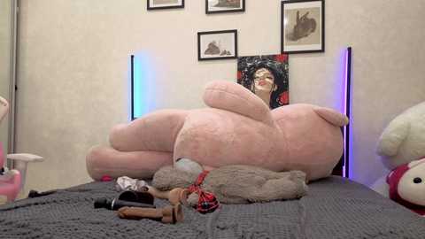 A video of a large pink teddy bear with an oversized head and floppy limbs, occupying a bed with grey sheets and plush toys, surrounded by framed art and a purple neon light.