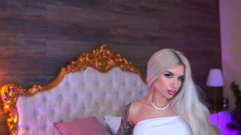 Video of a blonde woman with long hair, wearing a white off-shoulder dress and pearl necklace, posing seductively against a wooden wall with a golden, tufted headboard in a dimly-lit bedroom.