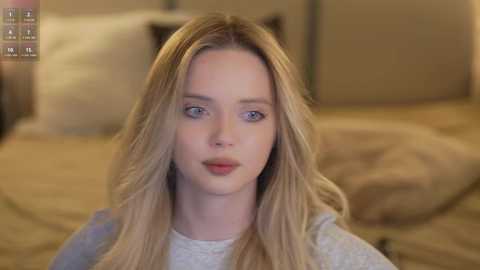 A video of a young woman with long blonde hair, light skin, blue eyes, and a neutral expression, wearing a grey sweater, sitting in a softly lit bedroom with beige bedding and pillows.
