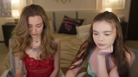 Video of two young women in a bedroom. One with long wavy hair, red lace lingerie, tattoos, and serious expression. The other with straight brown hair, green top, and pensive look.