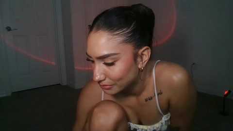 Video of a young woman with medium skin tone, dark hair in a bun, wearing a white spaghetti-strap top, sitting on a dark floor. She has closed eyes, and a tattoo on her left shoulder reads \"VIVI.\" The background features a gray door and a red light.