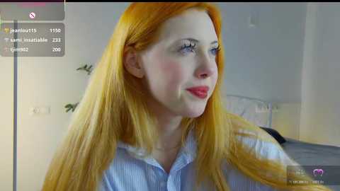 Video of a fair-skinned woman with long, straight orange hair, wearing a light blue button-up shirt, smiling slightly in a softly lit bedroom.