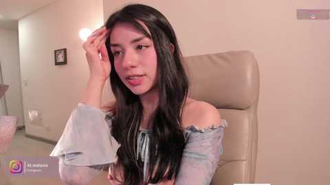 Video of a young woman with long black hair, wearing an off-shoulder light blue top, sitting in a beige leather chair in a modern room.