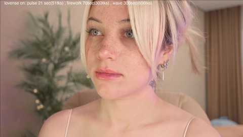 Media: Video of a young, fair-skinned woman with blonde hair in a messy bun, wearing light makeup and a beige top, against a blurred indoor background with a green plant.