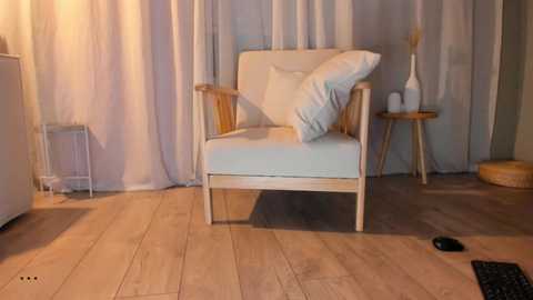 A video of a minimalist living room featuring a light wooden armchair with white cushions, a small round table, and beige curtains. The floor is light wood, and a black keyboard rests on the right.