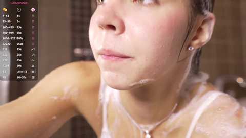 Video of a young woman with wet, slicked-back hair, wearing a white tank top, in a shower with soap suds on her face and chest.