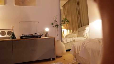 Video of a minimalist, warmly-lit bedroom with beige walls, a wooden floor, a white bed, a grey cabinet, a green plant, and a white vase.