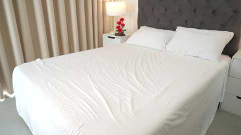 Video of a modern bedroom with a queen-sized bed covered in crisp white sheets and pillows, a grey tufted headboard, beige curtains, and a white nightstand with a red flower arrangement.