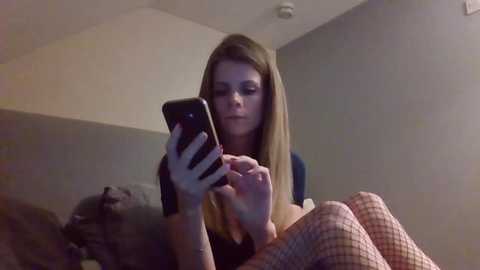 A video of a young woman with long, straight blonde hair, wearing a black top and red and white checkered leggings, sitting on a bed with a grey blanket, holding a black smartphone.