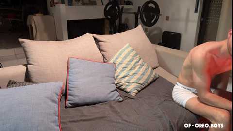 Video of a shirtless man with a white cast on his right arm, standing on a sofa with beige and blue cushions. Background shows a modern living room with a white wall, black bicycle, and a table.