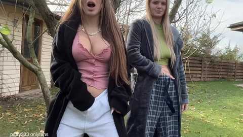 Video of two young women in casual, revealing outfits, one in a pink lace top and white pants, the other in a green crop top and plaid skirt, outdoors with a wooden fence and house in the background.